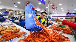 A worker in a seafood market dumps bright orange shrimp from a blue bin onto a bed of ice between a lot of other seafood laid out for sale. Other workers are visible in the background, as are the arms of shoppers placing orders from the other side of the ice bed.